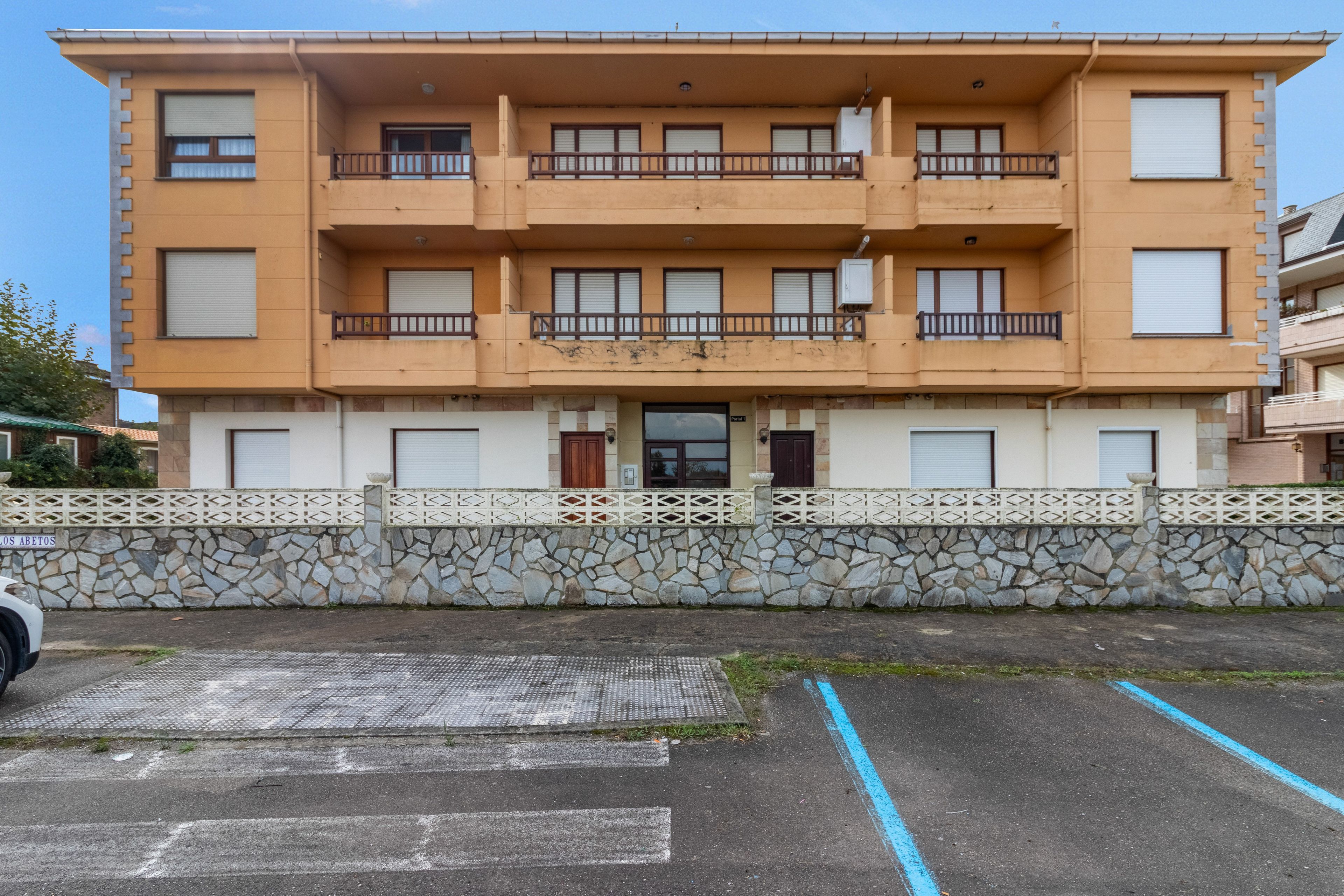 Un edificio residencial de varios pisos con balcones, una pared de piedra y una zona de estacionamiento con líneas azules.
