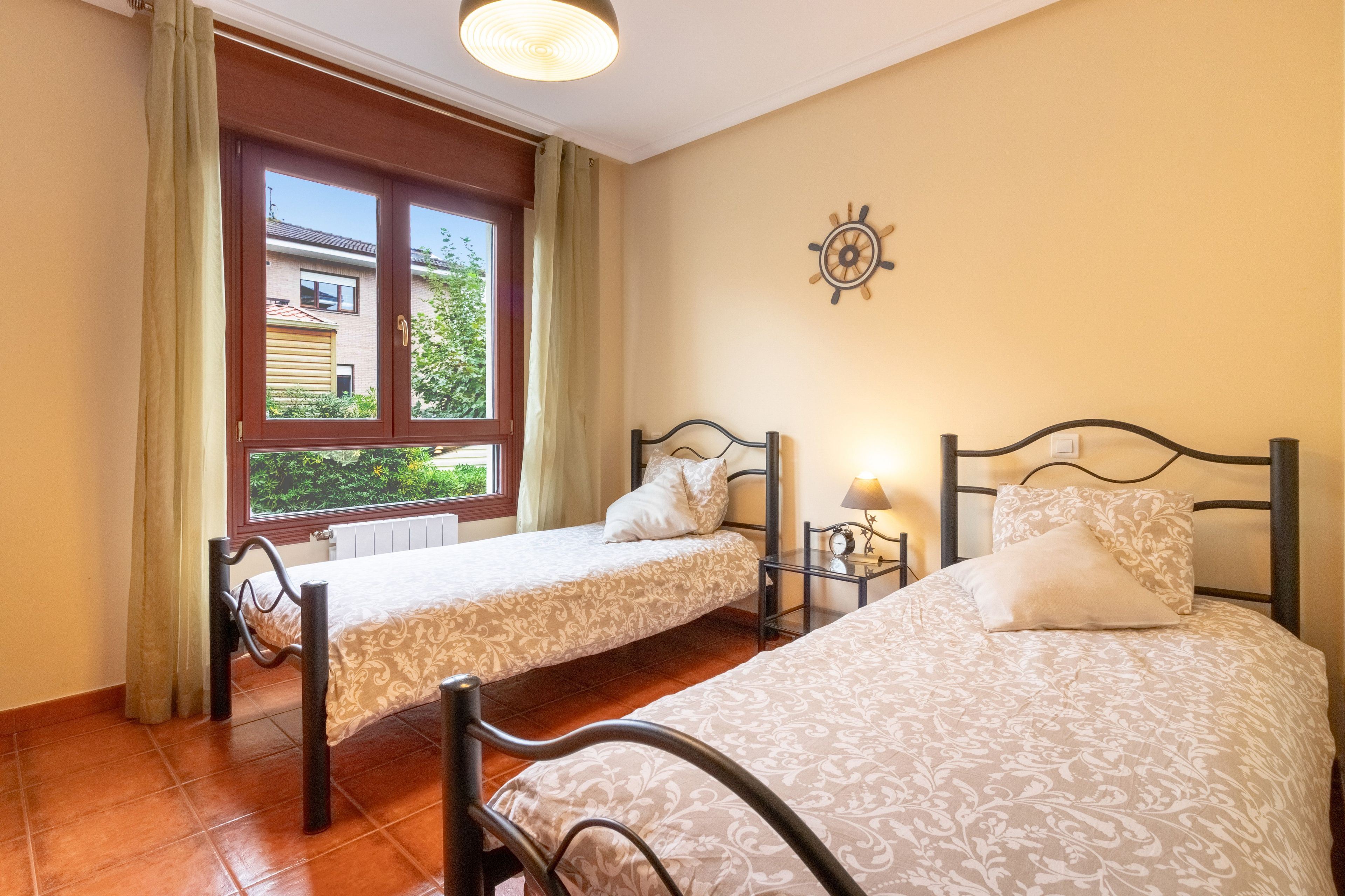 A room with two beds, a window with curtains, a wall clock, and a small table with a lamp.