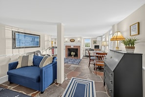 Ein Wohnzimmer mit blauem Sofa, Kamin, Essbereich mit Tisch und Stühlen sowie Küchenzeile mit Kühlschrank.