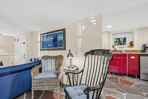 Ein Wohnzimmer mit einem blauen Sofa, Korbstühlen, einem Tisch mit einer Lampe, einem Gemälde eines Segelboots und einer Küchenzeile mit roten Schränken und einem Spülbecken.