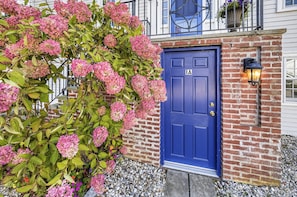 A blue door with the letter 'A' on it, a brick wall, and a hedge with pink flowers. - The Village Anchor/Cozy Flat near Harbor & Shops (Rockport)