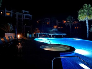 Uma área de piscina à noite, com espreguiçadeiras e uma banheira de hidromassagem, cercada por edifícios e palmeiras.
