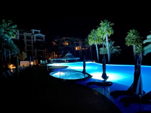 Área de piscina à noite com várias piscinas, palmeiras e um edifício ao fundo.