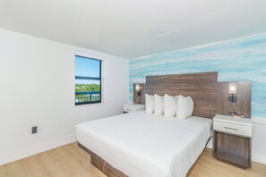 A bedroom with a large bed, a nightstand, a wall-mounted lamp, and a window with a view of greenery and a blue sky. - Myrtle Beach Oceanfront Retreat 2 BR Condo Yachtsman S706 (Myrtle Beach)