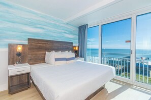 Ein Hotelzimmer mit einem großen Bett, einem Nachttisch, einer Lampe und Meerblick vom Balkon.