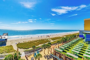 A beach view with a lifeguard tower, beach umbrellas, and a building with outdoor seating. - Myrtle Beach Oceanfront Retreat 2 BR Condo Yachtsman S706 (Myrtle Beach)
