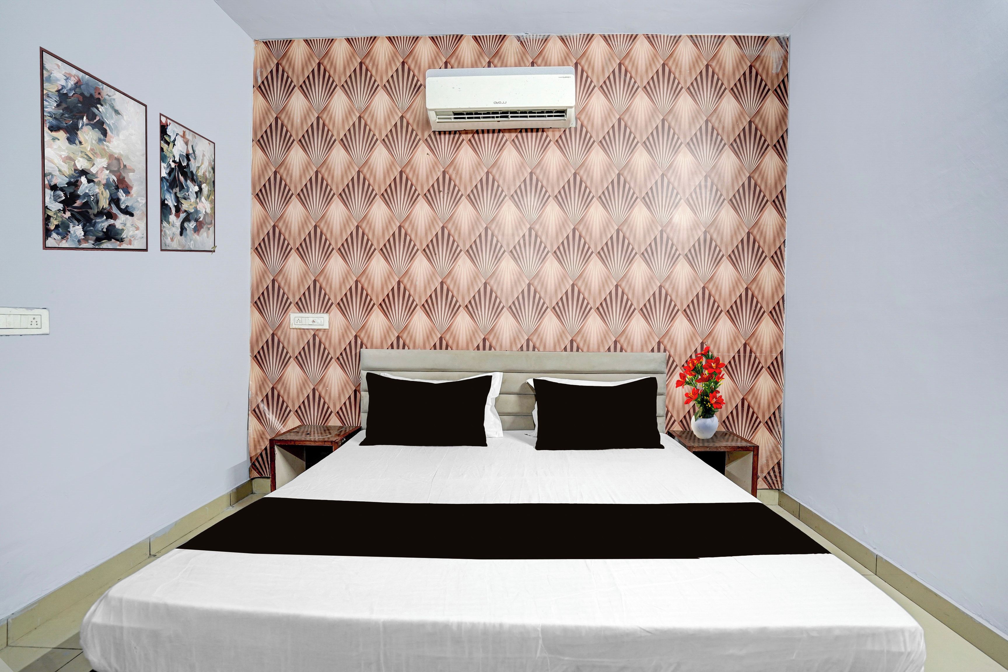 A hotel room with a bed, a nightstand, a vase with flowers, and an air conditioning unit.