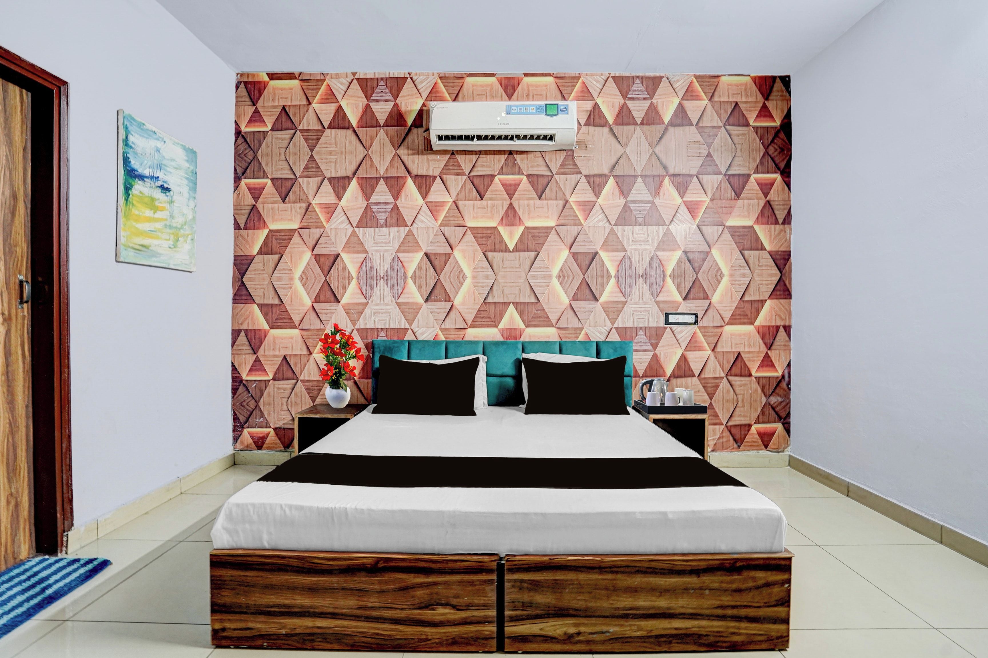 Un dormitorio con pared de diseño geométrico, cama con ropa de cama blanco y negro, cabecera color turquesa, mesita de noche con lámpara y una planta en maceta.