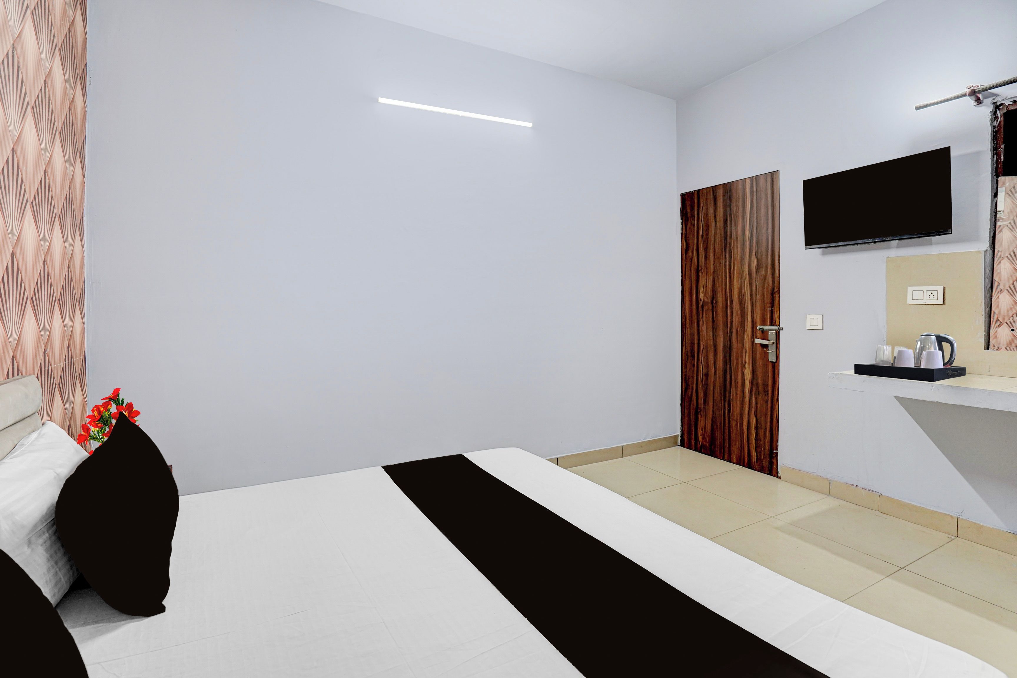 Una habitación de hotel con una cama, un televisor montado en la pared, una puerta de madera y una mesita con una tetera y tazas.