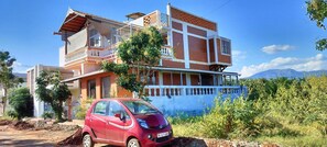A two-story building with a balcony, a red car parked in front, and a mountainous landscape in the background. - Eco-friendly 2-bedroom house with mountain view in charming Thenkarai with AC (Thenkarai)