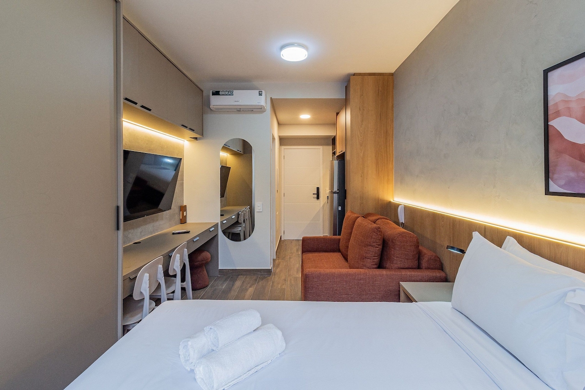 Quarto de hotel com cama, televisão, sofá e pequena área de refeições.