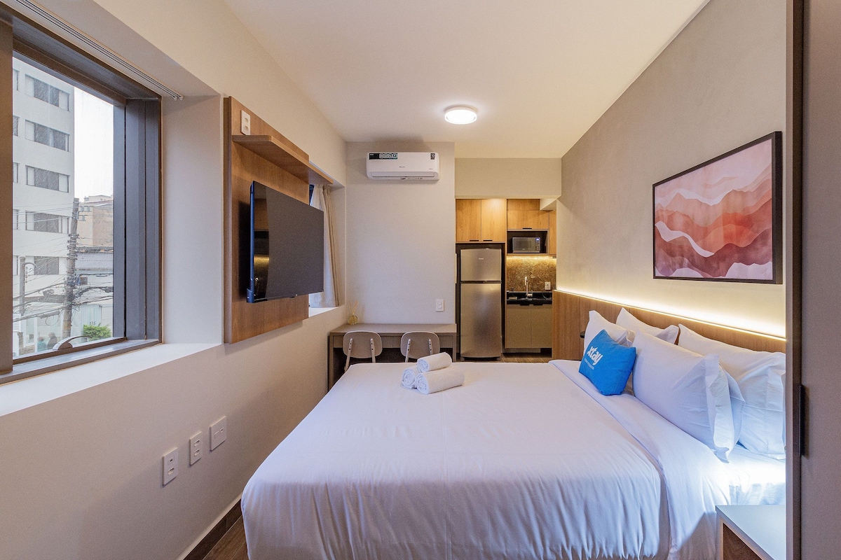Quarto de hotel com cama, escrivaninha, TV de tela plana na parede, micro-ondas, geladeira e ar-condicionado.