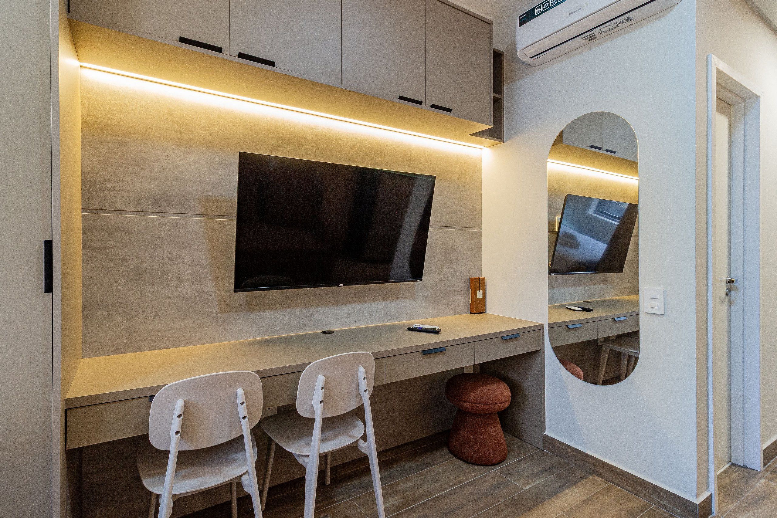 Een moderne kamer met een flatscreen televisie aan de muur, een bureau met twee stoelen en een ronde spiegel.