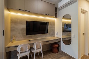 Una habitación moderna con televisión de pantalla plana en la pared, un escritorio pequeño con dos sillas y un espejo redondo.