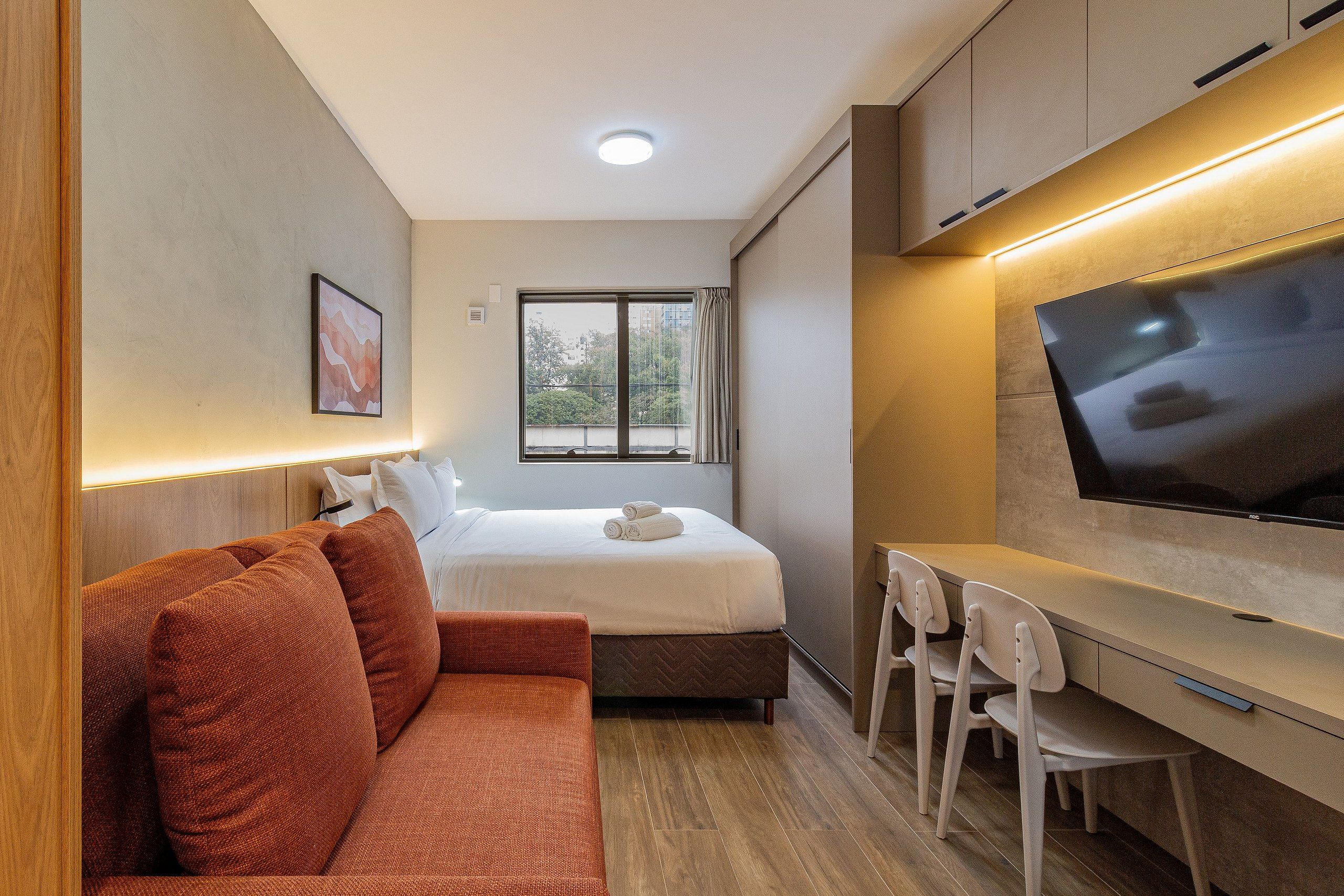 Een kleine hotelkamer met een bed, een slaapbank, een bureau met stoelen, een televisie en een raam met gordijnen.