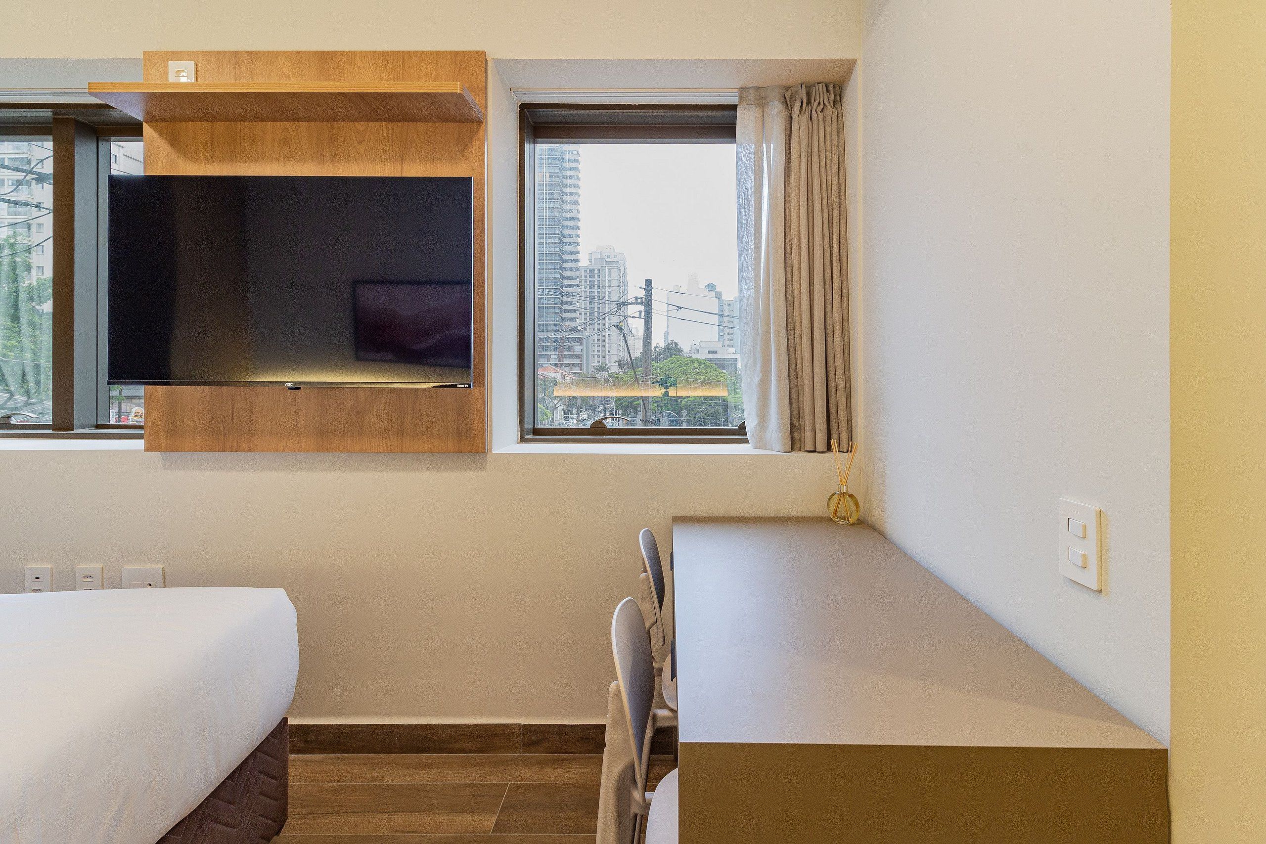 A hotel room with a bed, a desk, a chair, a window with a view of buildings, and a shelf with a TV.