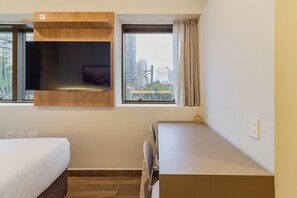 A hotel room with a bed, a desk, a chair, a window with a view of buildings, and a shelf with a TV. - Xtay Villa Minerva (São Paulo)
