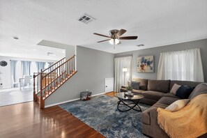 A modern living room with a wooden staircase, a ceiling fan, a round coffee table, and a large window with curtains. - 3BR Home Near Las Vegas Strip I Mid-Term Stays (Las Vegas)