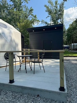 A covered outdoor seating area with a table and chairs, a small building, and a gravel path.