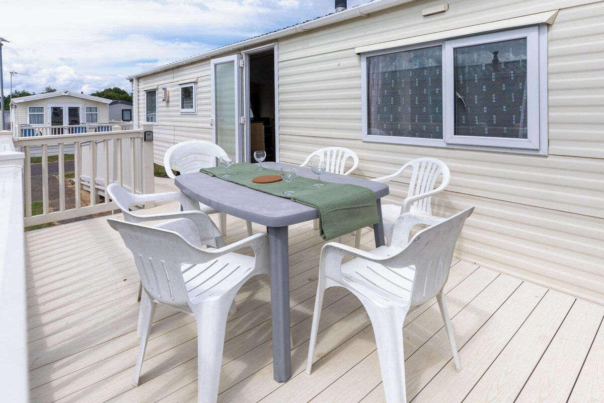 Une maison mobile dotée d’une terrasse, d’une table avec des chaises et d’une fenêtre avec des rideaux.