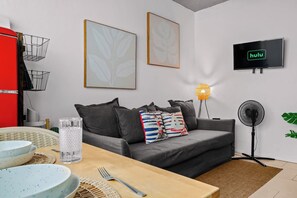 A modern living room with a grey sofa, a dining table with plates and cutlery, a red appliance, two framed artworks, a wall-mounted TV, and a standing fan. - Casa Esmeralda – Old San Juan (San Juan)