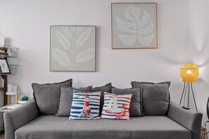 Un canapé gris agrémenté de deux coussins à motifs, un tableau représentant une feuille encadrée, une lampe sur pied et une bibliothèque contenant des livres et des objets décoratifs.
