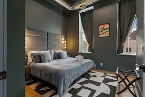 A modern bedroom with a large bed, a headboard with lights, a window with curtains, and a wall-mounted poster. - Lofted Luxury | 5 BR • Sleeps 10 • Downtown! (Frankfort)