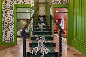Un escalier aux murs verts et au papier peint à motifs, menant à une pièce où se trouvent un lit et une fenêtre.