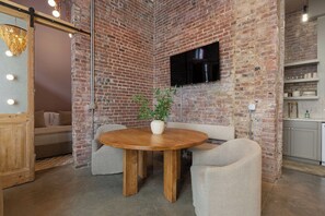 A room with a brick wall, a round wooden table with chairs, a flat-screen TV mounted on the wall, and a small plant on the table. - Suite Luxury | Designer • Ideal Downtown Location! (Frankfort)