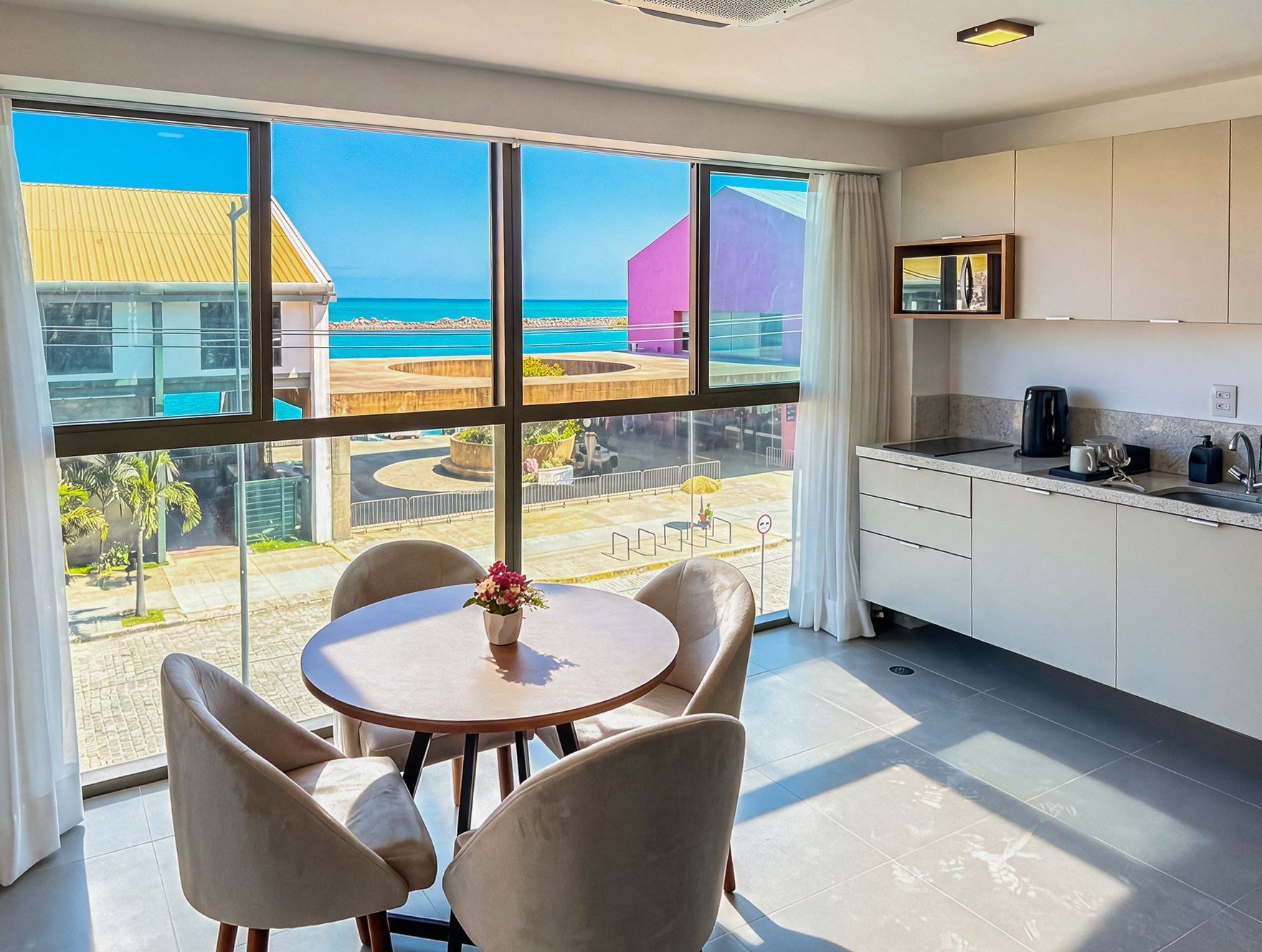 Une chambre moderne dotée d’une grande fenêtre donnant sur une piscine et des immeubles, d’une table ronde avec des chaises et d’un coin cuisine.