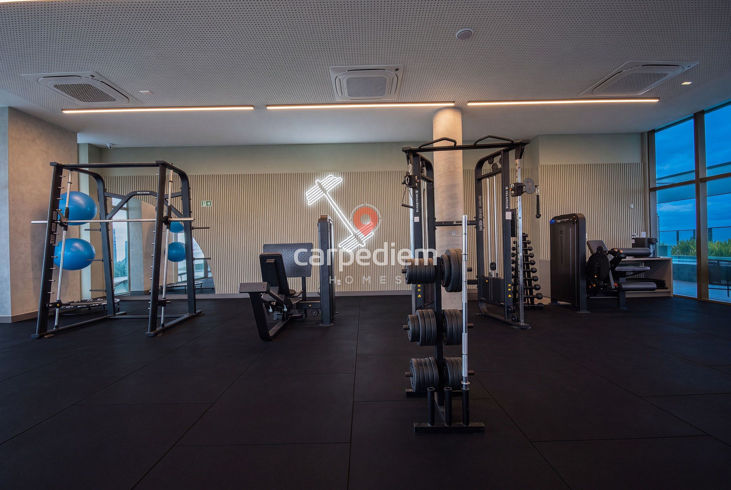 A modern gym with various exercise equipment, including weight machines and free weights, and a large window providing a view of the outdoors.