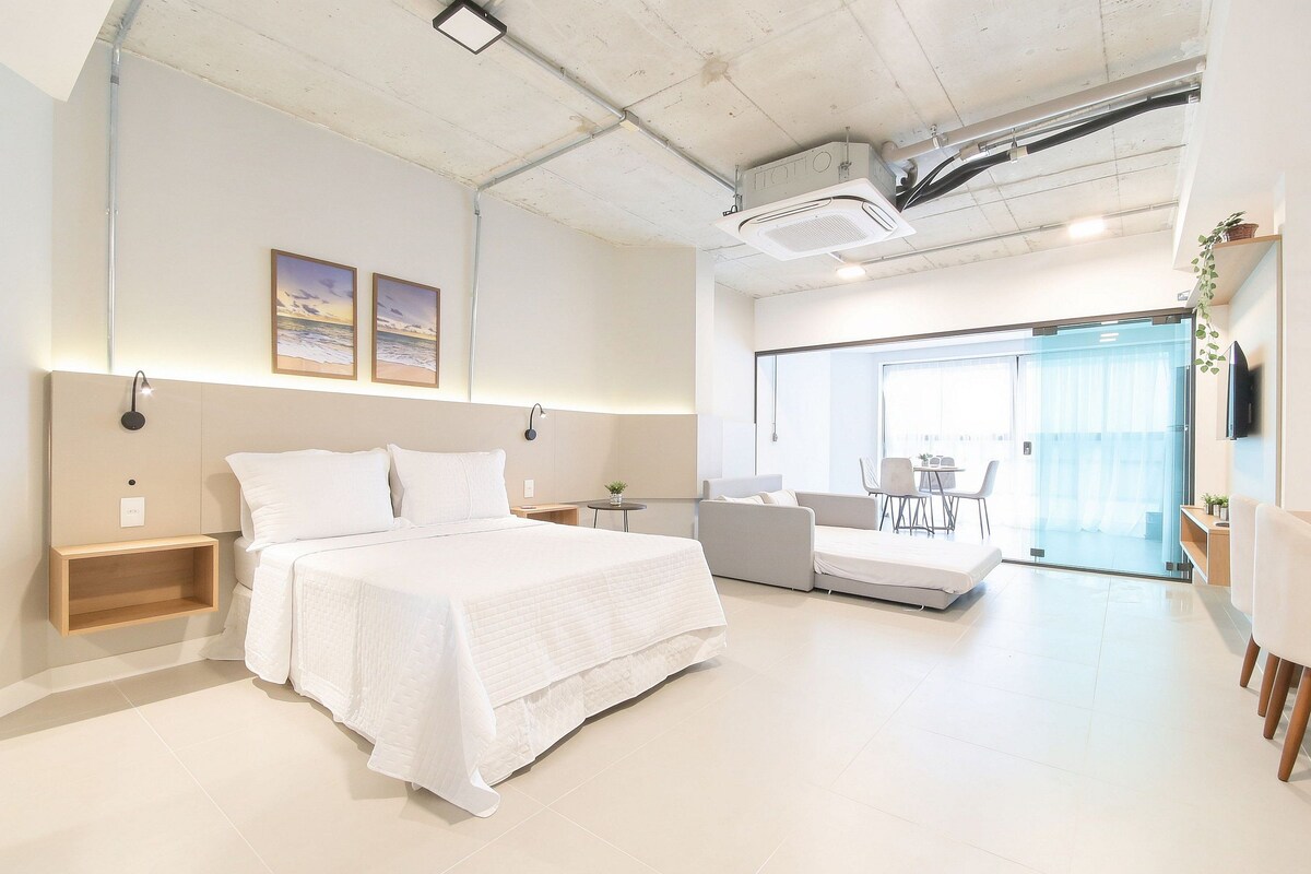 Quarto de hotel moderno com uma cama grande, um sofá, uma mesinha e uma televisão.