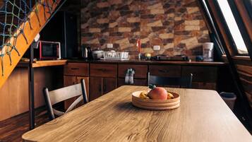 A wooden dining table with a fruit bowl, a chair, and a kitchen in the background with a stone wall.