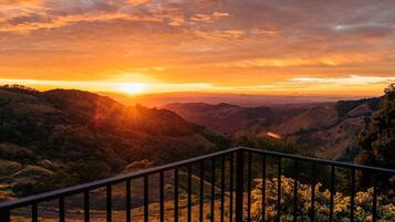 A sunset view from a balcony with a railing, overlooking a valley with hills and trees.