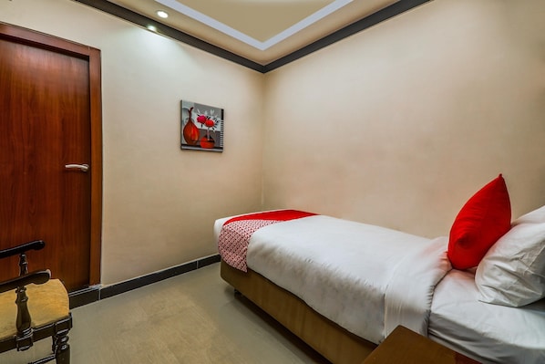 Una habitación de hotel con puerta de madera, una cama con ropa blanca y almohadas rojas, una mesita con una silla y un cuadro en la pared.
