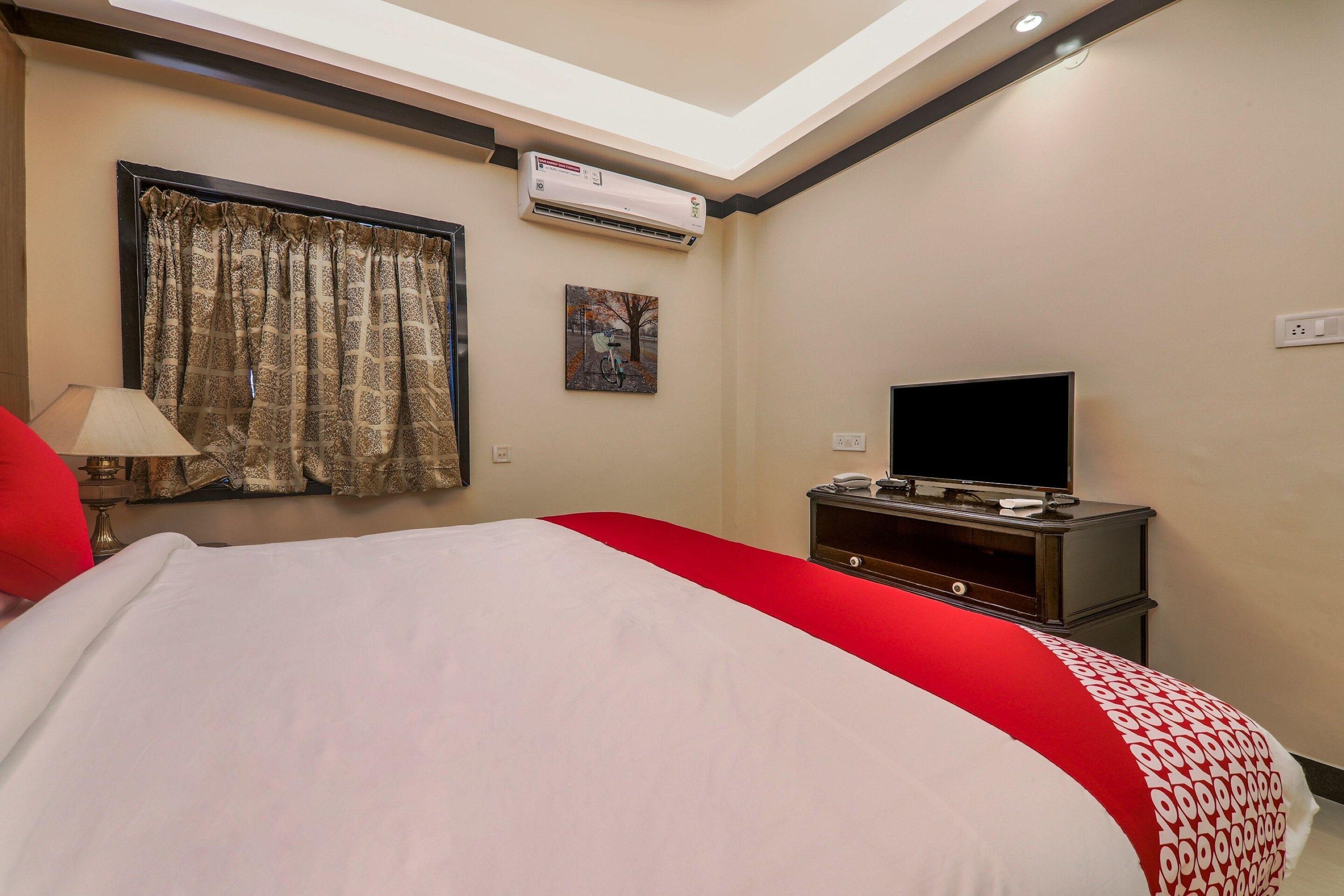 Habitación de hotel con cama, televisor, equipo de aire acondicionado, ventana con cortinas y un cuadro en la pared.