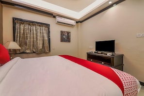 Habitación de hotel con cama, televisor, equipo de aire acondicionado, ventana con cortinas y un cuadro en la pared.