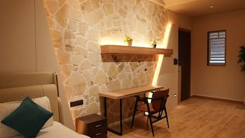 A bedroom with a stone wall, a bed with a teal pillow, a desk with a chair, and a ceiling fan.