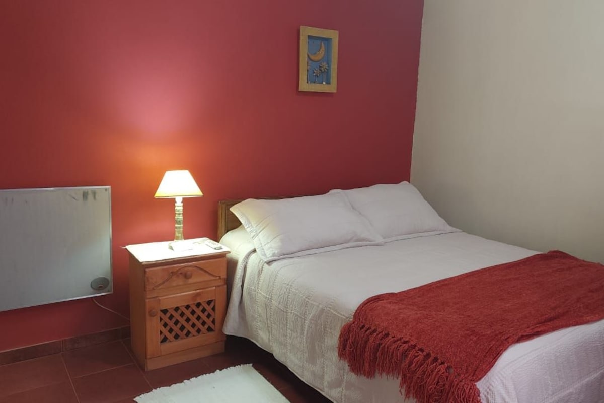 Un dormitorio con una cama, una mesita de noche, una lámpara, un cuadro en la pared y una manta roja.
