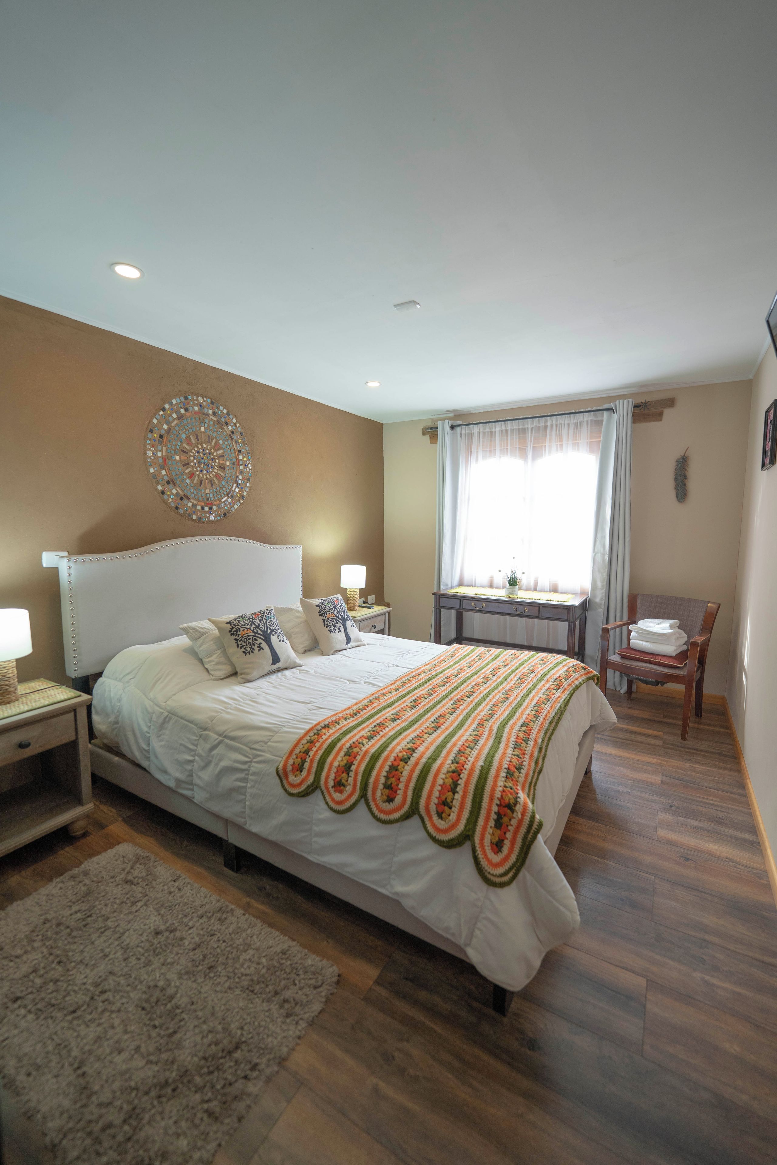 A bedroom with a large bed, a bedside table, a nightstand, a window with curtains, a wooden chair, and a decorative wall piece.