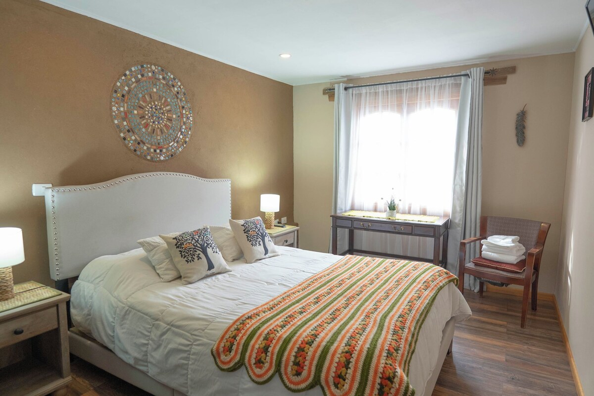 A bedroom with a large bed, a bedside table, a nightstand, a window with curtains, a wooden chair, and a decorative wall piece.