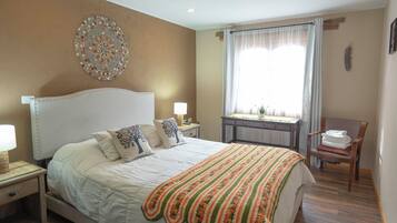 A bedroom with a large bed, a bedside table, a nightstand, a window with curtains, a wooden chair, and a decorative wall piece.