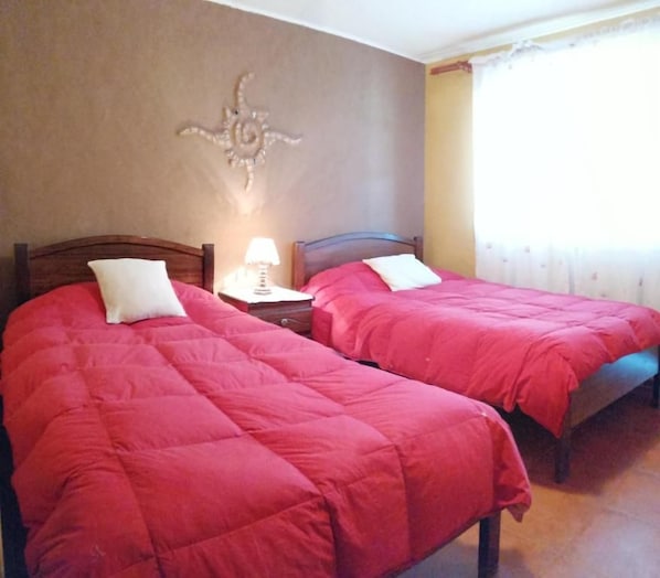 A room with two beds, each with a red bedspread and white pillows, a nightstand with a lamp, and a decorative wall piece.