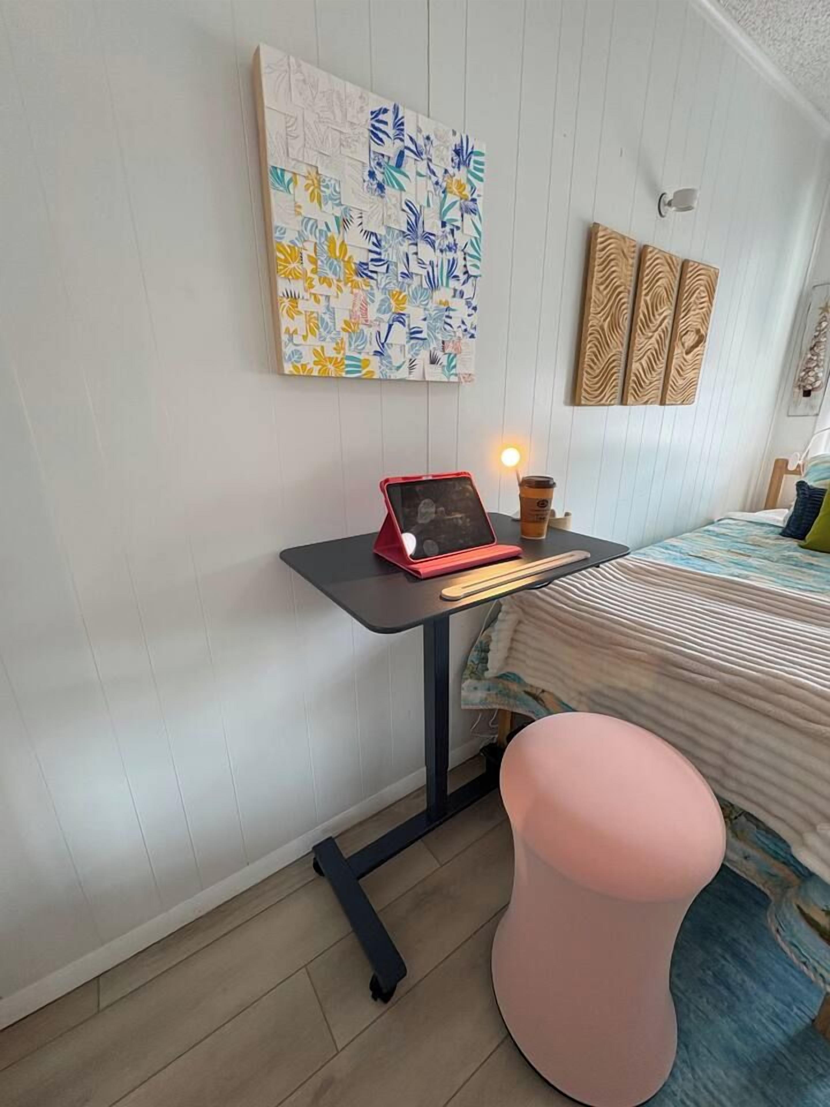 A room with a bed, a desk with a laptop, a pink stool, and a wall-mounted artwork.