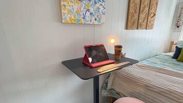 A room with a bed, a desk with a laptop, a pink stool, and a wall-mounted artwork.