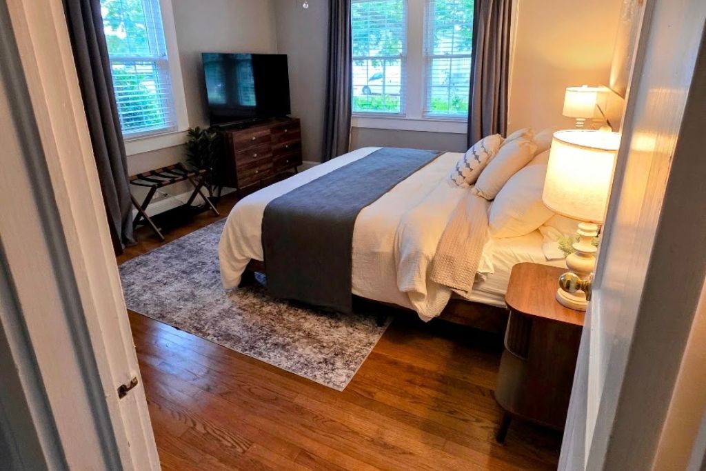 A bedroom with a bed, bedside table, lamp, TV, dresser, and windows.