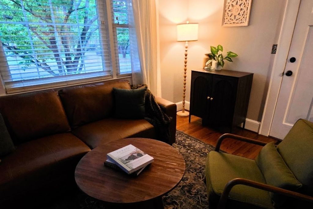 Un salon avec un canapé en cuir marron, une table basse ronde sur laquelle se trouve un magazine, un fauteuil vert, un meuble en bois foncé et un lampadaire.