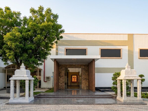 Entrada de um edifício com caminho de mármore, duas colunas brancas e uma grande árvore.