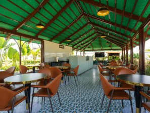 Uma área de estar coberta ao ar livre, com mesas e cadeiras, cozinha e vista para o verde.