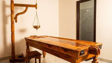 A wooden massage table with a footrest and a wooden chair, a hanging brass scale, and a marble-tiled floor.